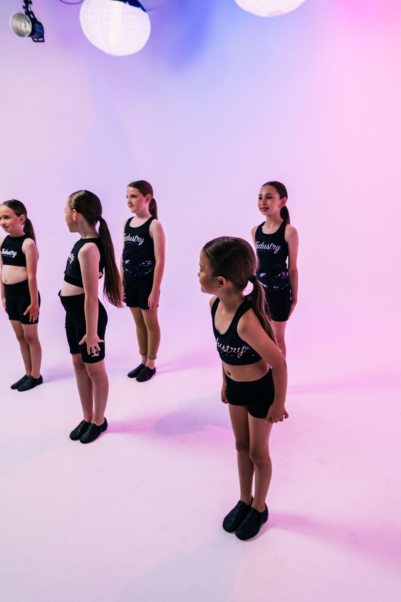 A line of young dancers in black posing on a pink backdrop