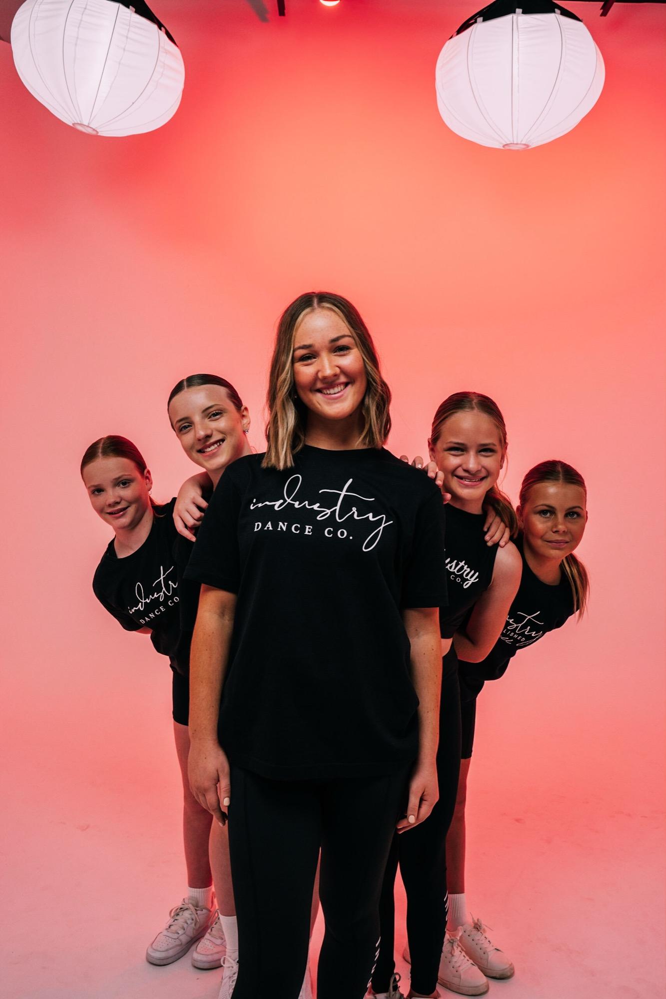 Senior dancers grouped closely with their teacher against a coral backdrop