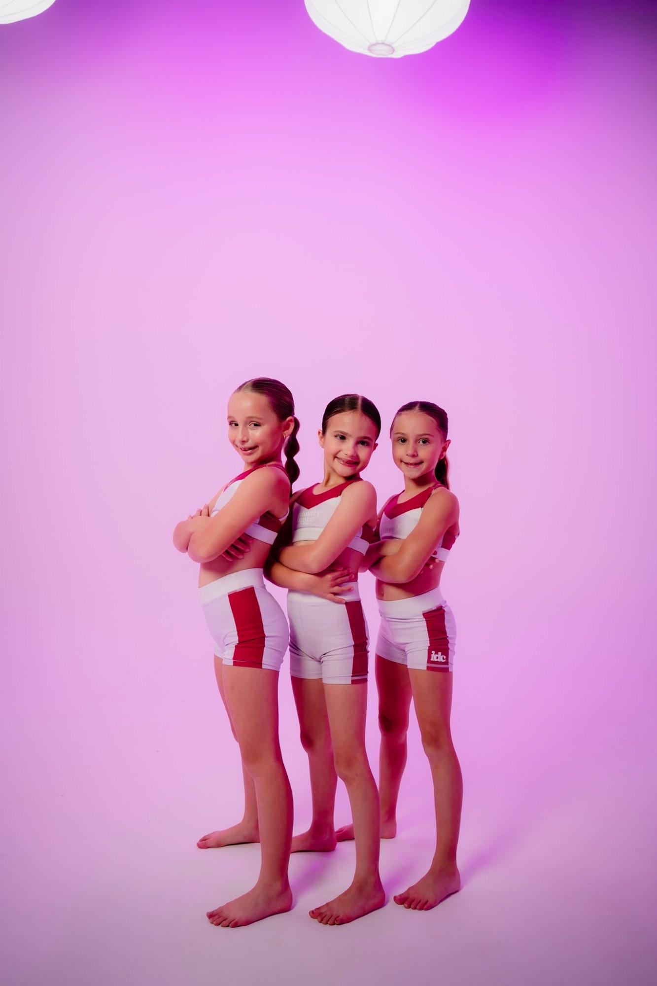 Three Industry Dance Co. dancers in IDC uniforms, arms crossed and smiling, against a pink studio backdrop